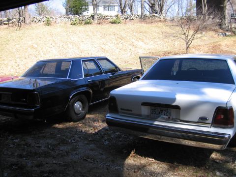 1991 Ford LTD Crown Victoria - Two Ford Crown Victorias, one black and one white, are parked on a dirt lot with a stone wall and house in the background.