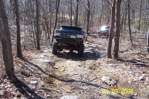 1979 Ford Bronco - A black SUV navigates a challenging rocky trail through a wooded area, with another vehicle following behind.