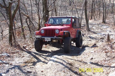 1979 Ford Bronco - A red Jeep Wrangler navigates a challenging off-road path through a wooded area.