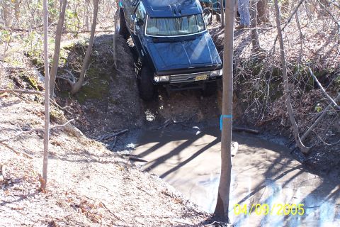 1979 Ford Bronco - A black SUV navigates a challenging off-road trail, descending into a muddy water obstacle.