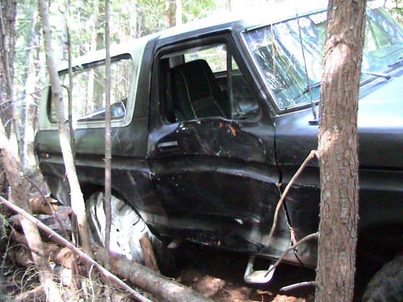 Black 1978 Ford Bronco with a white top, damaged on the driver's side, partially obscured by trees and logs