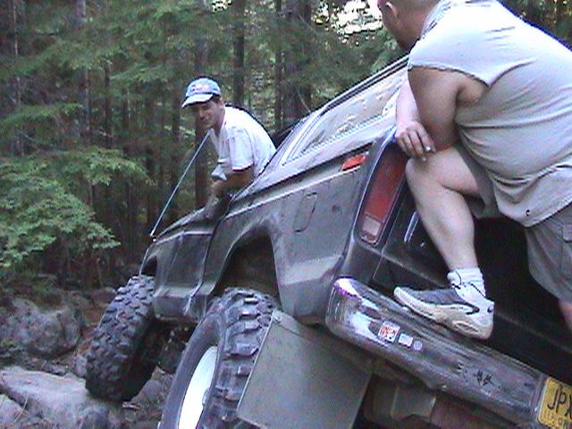 Dark 1978 Ford Bronco off-roading, front passenger side angled up on rocks, driver leaning out window