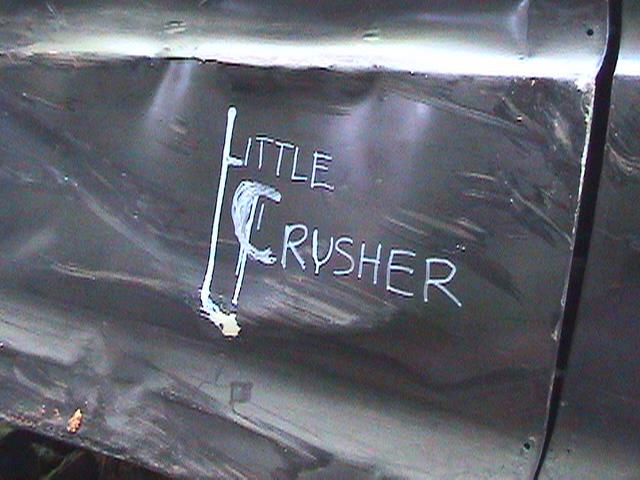 Dark-colored vehicle side panel with white text 'LITTLE CRUSHER' and visible dents and scratches