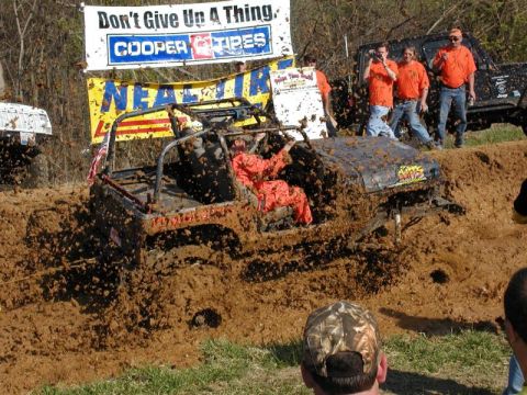 1992 Ford Bronco - A driver in an orange suit navigates an off-road vehicle through a deep mud pit, with spectators watching.
