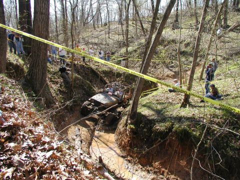 1992 Ford Bronco - Spectators watch an off-road vehicle navigate a challenging muddy course in a wooded area.
