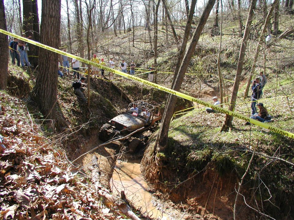 Off-road vehicle with roll cage driving through a muddy, wooded ravine with spectators watching