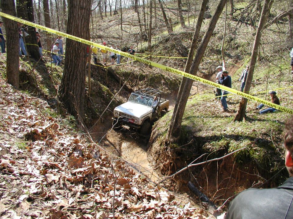 White 1970s off-road SUV driving through a muddy ravine with caution tape overhead