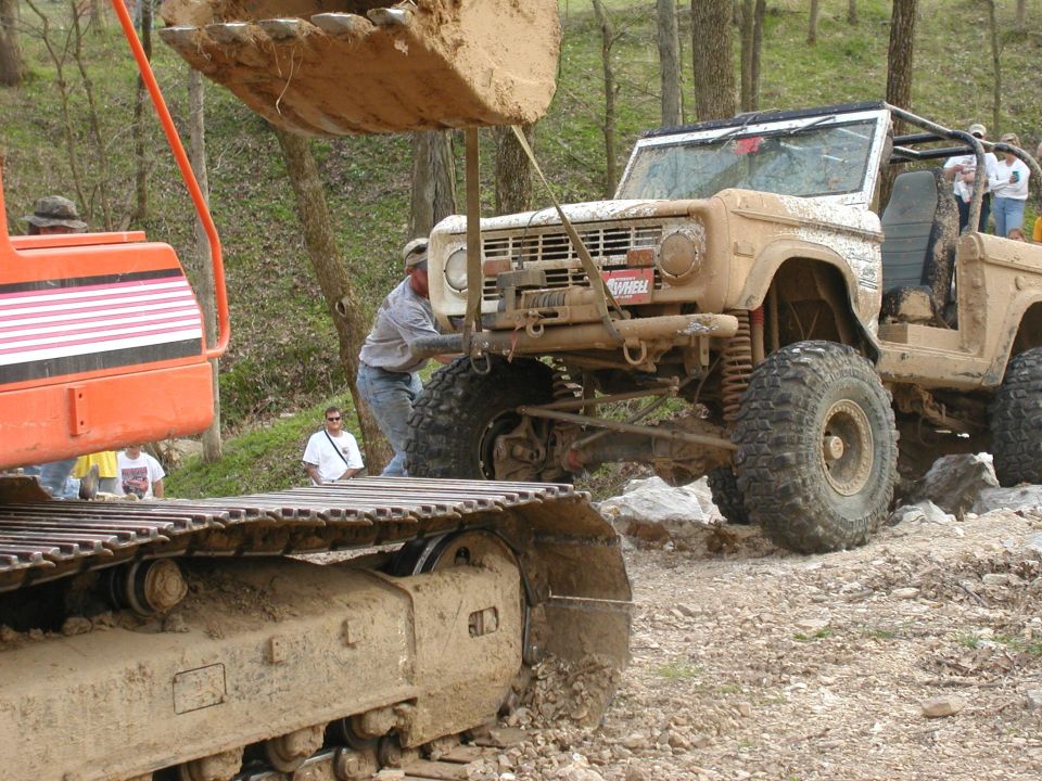 Muddy off-road SUV with roll cage being lifted by excavator bucket