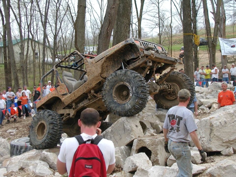 Muddy off-road vehicle climbing large rocks, with spectators watching