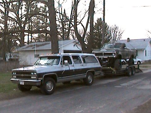 1977 Ford F-150 - A two-tone Chevrolet Suburban tows a lifted pickup truck on a trailer down a residential street.