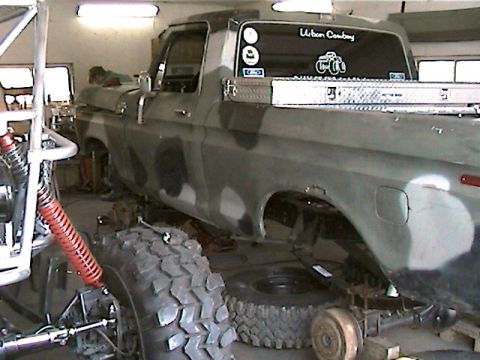 1977 Ford F-150 - A 1977 Ford F-150 pickup truck with camouflage paint is shown in a garage, undergoing modifications with large off-road tires.