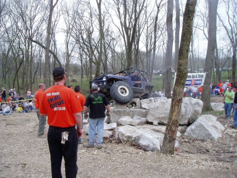 1992 Ford Bronco - An off-road vehicle attempts to climb over a large rock obstacle with a crowd of spectators gathered around.
