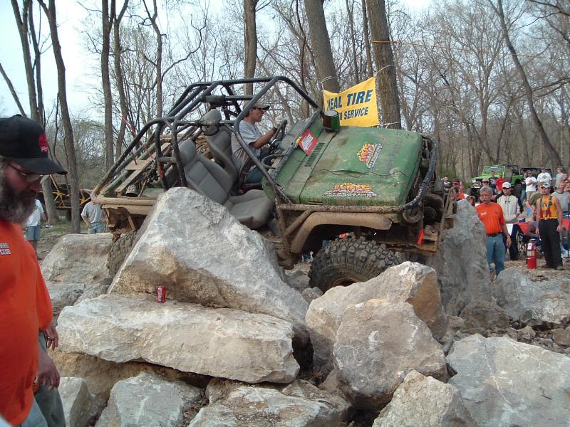 Off-road buggy with roll cage climbing over large rocks, crowd watching. Yellow banner in background