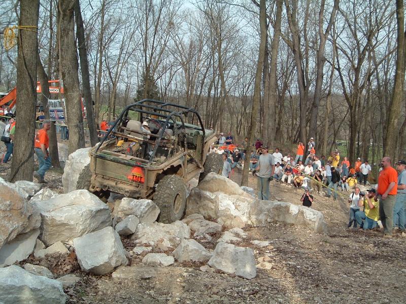 Off-road vehicle with roll cage climbing over large rocks, spectators watch from hillside