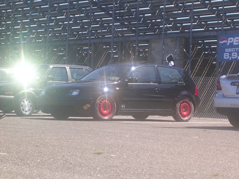 Black hatchback with red rims parked next to another car, with stadium seating in the background