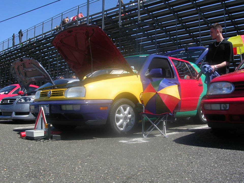 Multi-colored Volkswagen Golf with hoods open, parked in front of bleachers