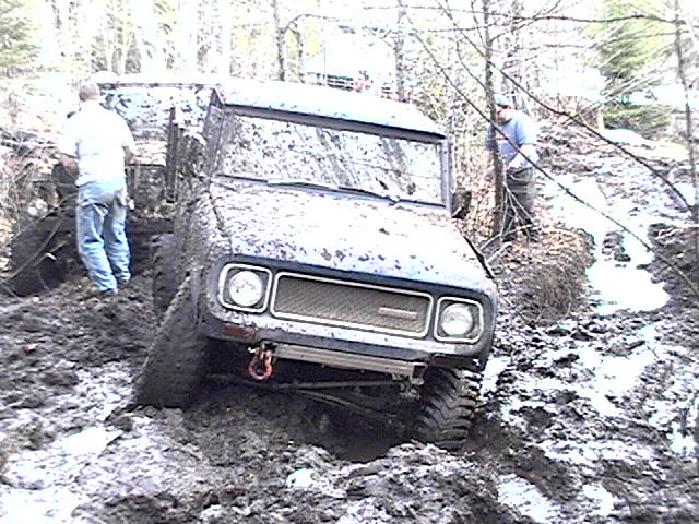 Mud-covered International Scout stuck deep in a mud pit with two people nearby in the woods