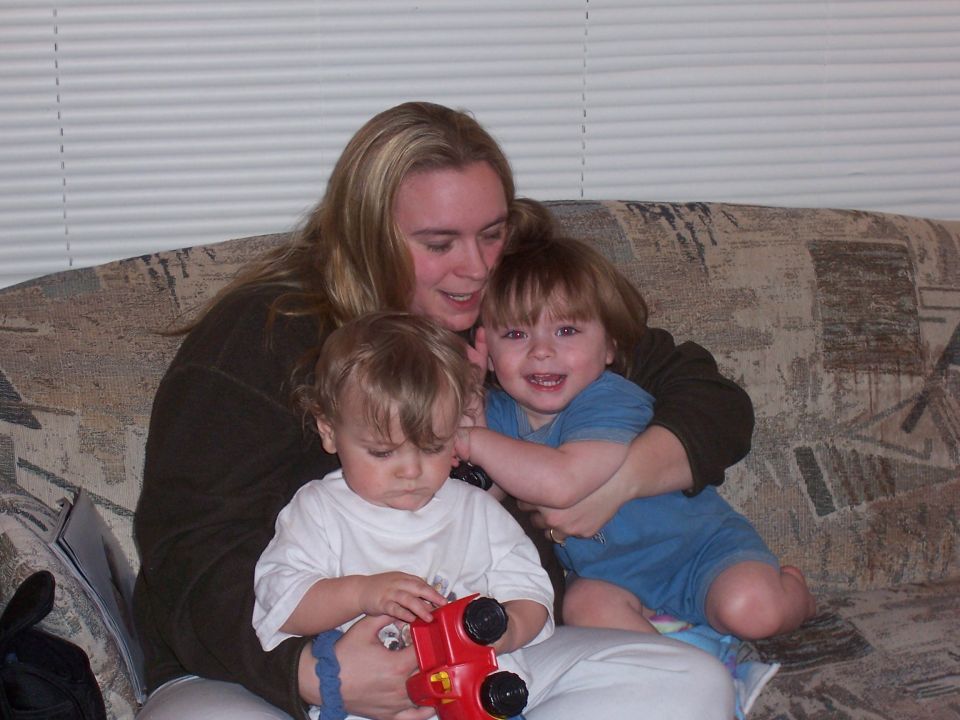 Woman holding two young children on a couch, one child plays with a red toy truck