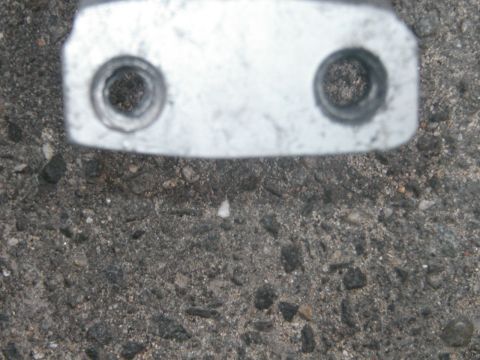 1994 Ford Bronco - A close-up view of a metal bracket with two bolt holes, resting on a textured asphalt surface.