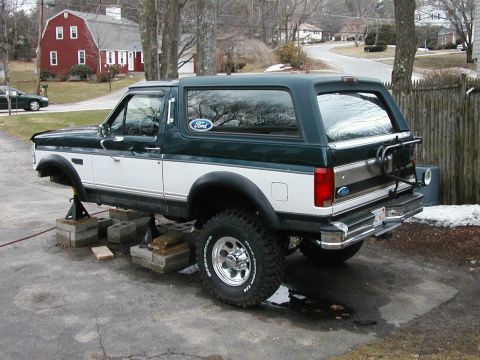1994 Ford Bronco - A two-tone Ford Bronco is raised on jack stands in a driveway, undergoing maintenance.