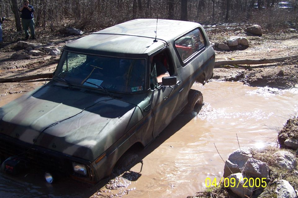 Camo-painted Ford Bronco crossing muddy water on an off-road trail