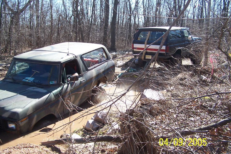 Camo-painted Ford Bronco stuck in mud being pulled by a white and blue Bronco on a wooded trail