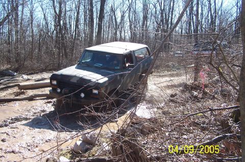 1979 Ford Bronco - A dark SUV is seen off-roading through a muddy and wooded area, with other vehicles visible in the background.