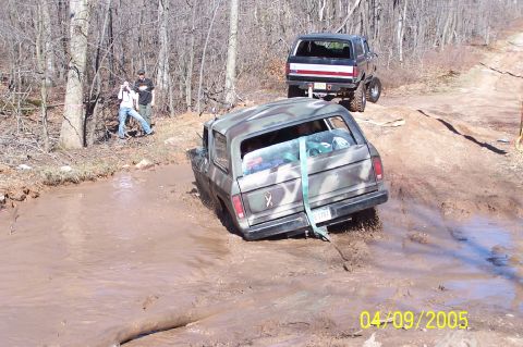 1979 Ford Bronco - Two SUVs navigate a muddy off-road trail with trees in the background.
