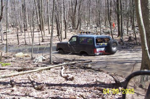 1979 Ford Bronco - A dark blue 1979 Ford Bronco navigates a muddy trail in a forest, with its rear spare tire prominent.