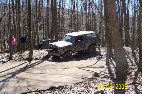1979 Ford Bronco - Two off-road vehicles are seen navigating a muddy trail in a wooded area.