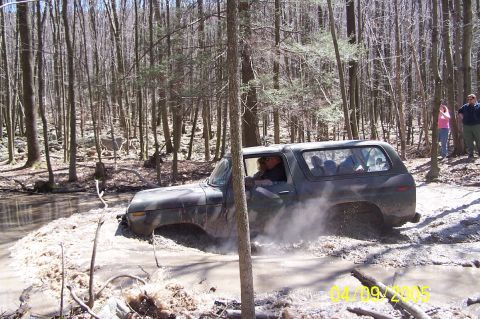 1979 Ford Bronco - A Ford Bronco navigates a muddy trail, kicking up water and dirt as it goes through the wooded terrain.