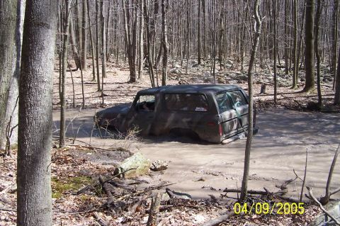 1979 Ford Bronco - A camouflaged Ford Bronco navigates a muddy forest trail, partially submerged in water.