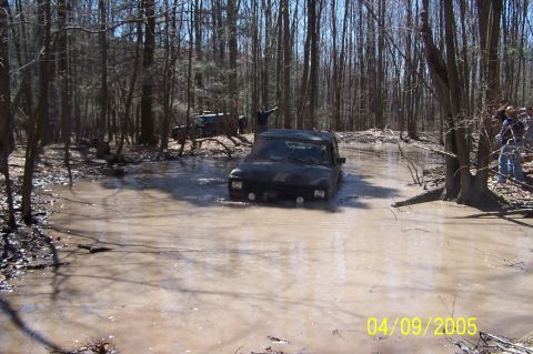 1979 Ford Bronco - Off-road vehicles tackle a challenging muddy water obstacle in a wooded area.