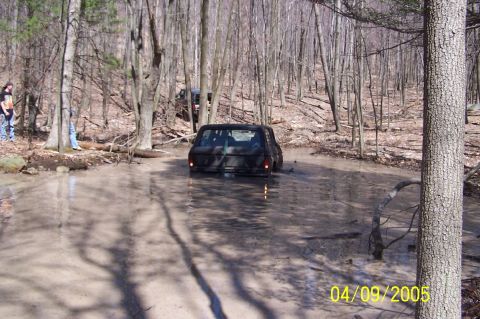 1979 Ford Bronco - A black SUV navigates a muddy trail in a wooded area, with another vehicle visible in the background.
