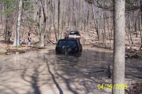 1979 Ford Bronco - Two SUVs navigate a muddy trail through a forest, with one vehicle deeply mired in the muck.