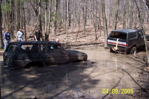 1979 Ford Bronco - Two SUVs are stuck in the mud in a wooded area with people observing.