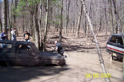 1979 Ford Bronco - Two off-road vehicles are stuck in a muddy puddle in a forest with several people observing.