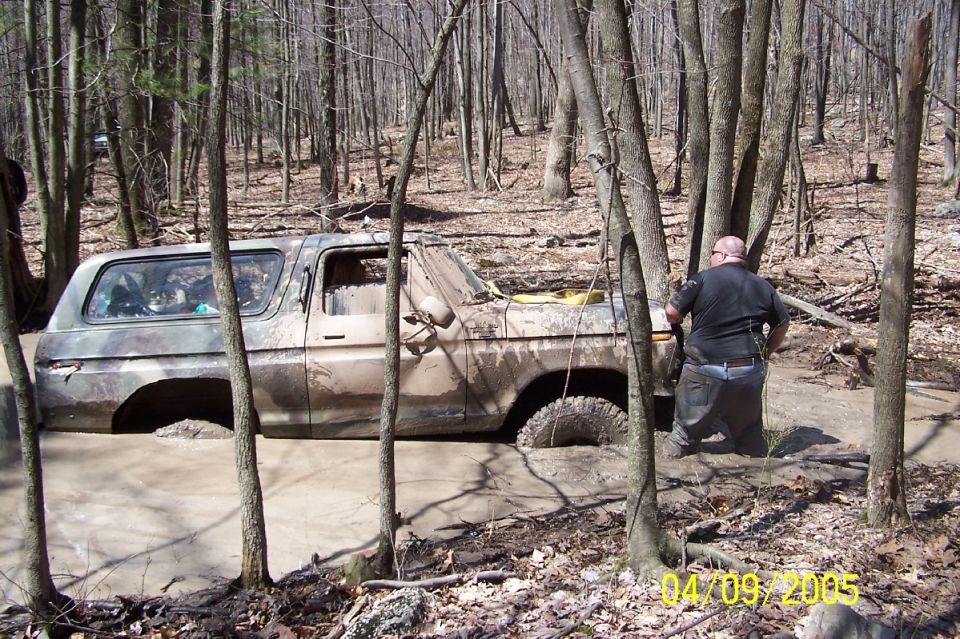 Muddy 1978 Ford Bronco stuck in a wooded area, with a person nearby