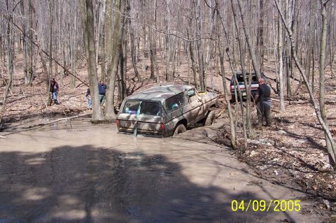 1979 Ford Bronco - Two off-road vehicles are in a wooded area, with one deeply stuck in a muddy creek.