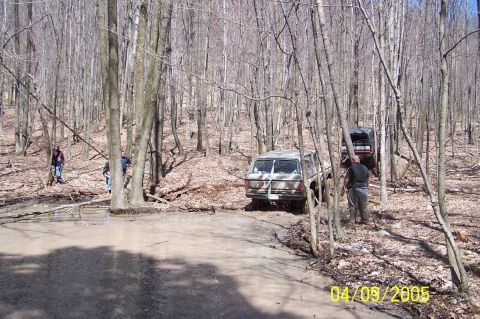 1979 Ford Bronco - Two SUVs navigate a wooded area with muddy terrain and sparse trees.