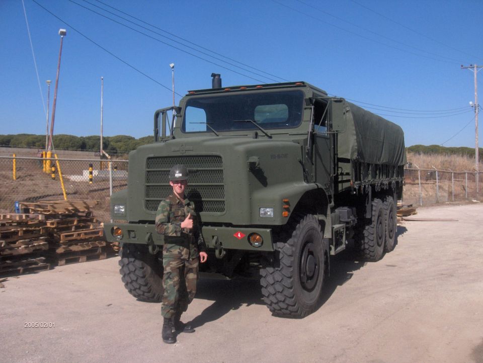 Green military truck with a person in camouflage standing in front. The truck has a tarped cargo bed