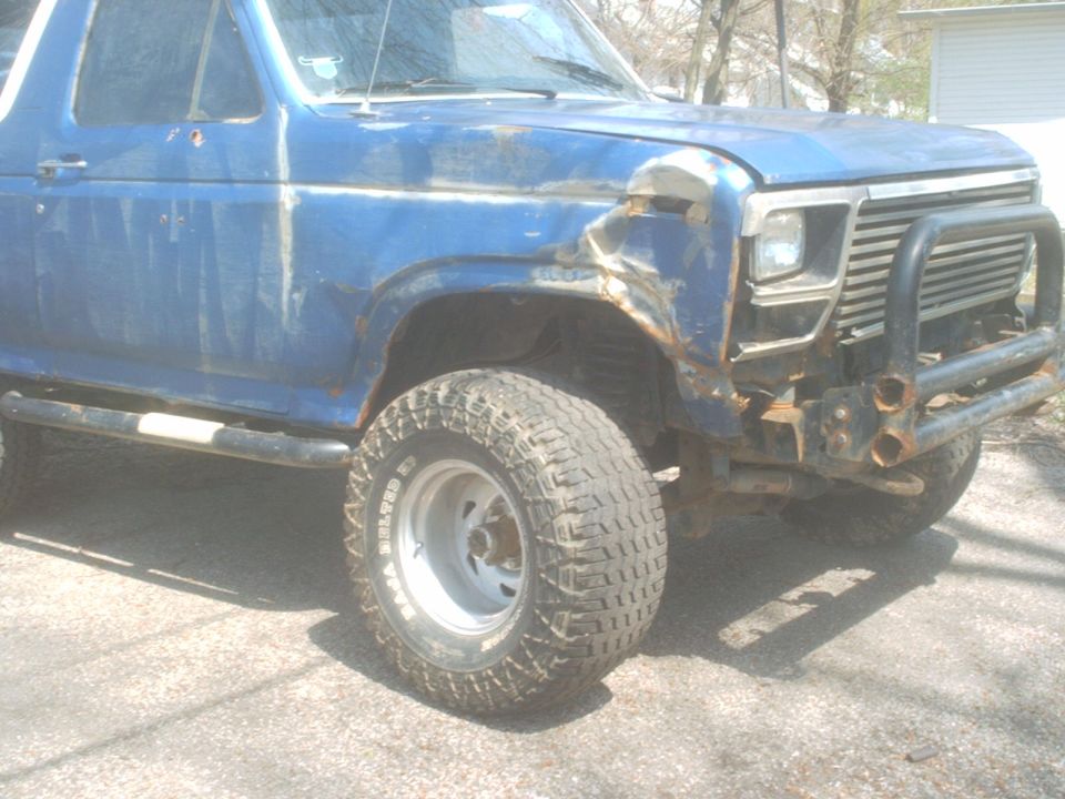 Blue Ford Bronco with a dented fender and aftermarket bumper. It has large off-road tires