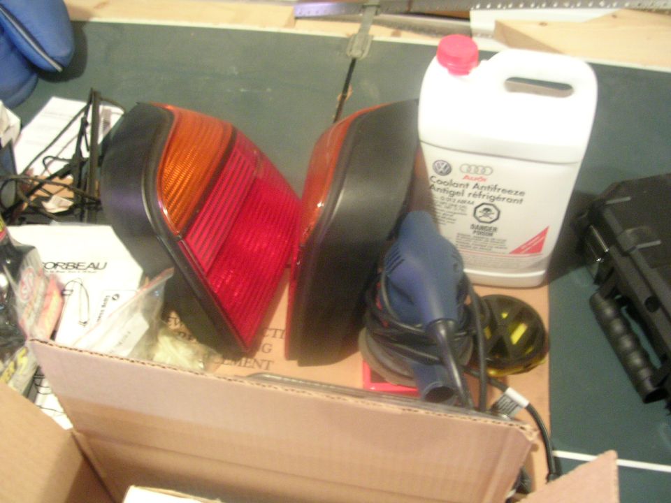 Pair of red and amber tail lights in a box next to a coolant jug and orbital sander