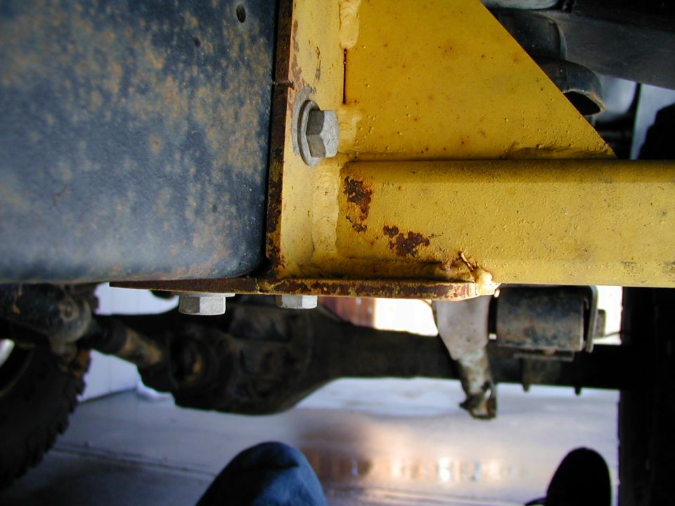 Close-up of yellow painted metal bracket bolted to a dark blue frame rail, with suspension parts and differential visible below
