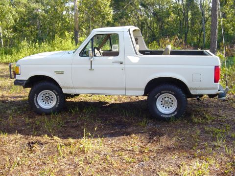1989 Ford Bronco - This white Ford Bronco is parked outdoors in a grassy area with trees. The vehicle is shown from the side.