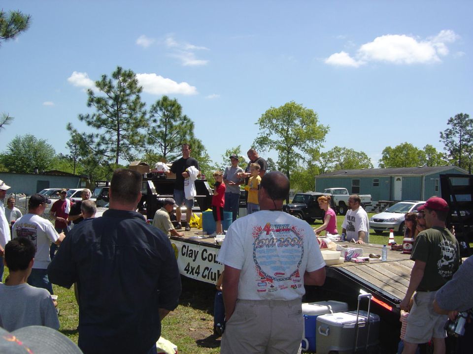People gathered outdoors for an event, with several vehicles visible in the background and on a trailer