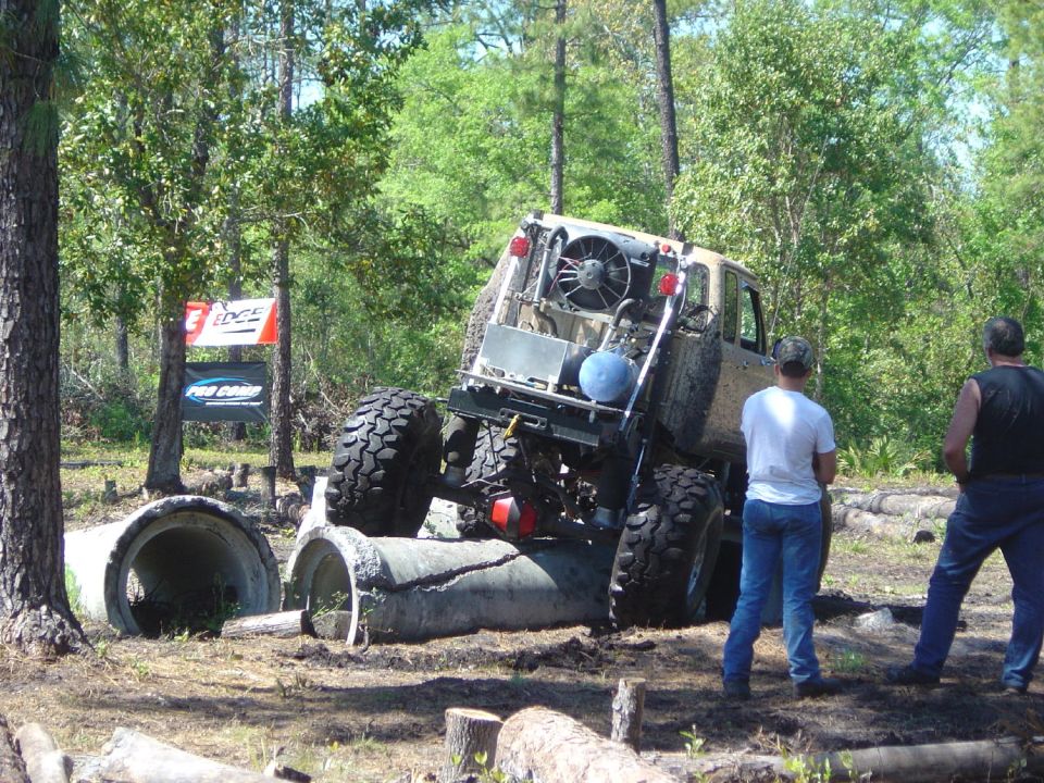 Mud-splattered off-road vehicle with large tires, rear-mounted fan, and custom roll cage, navigating concrete pipes