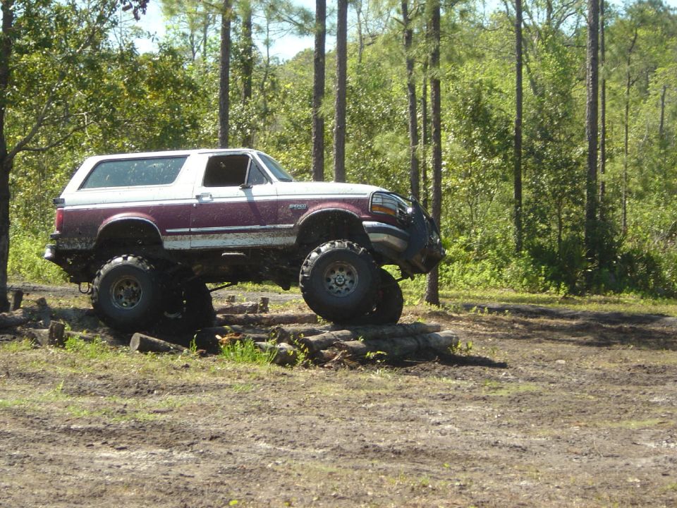 1993 Ford Bronco with large tires driving over logs in a wooded area
