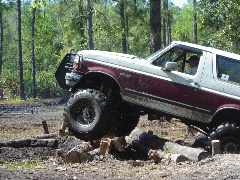 Two-tone maroon and white Ford Bronco with lifted suspension and large tires navigating an obstacle course of logs