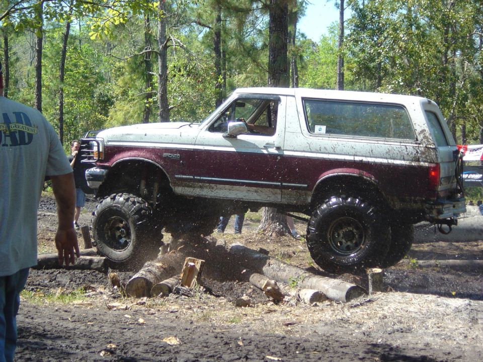 1993 Ford Bronco with lifted suspension and large tires driving over logs on a muddy trail